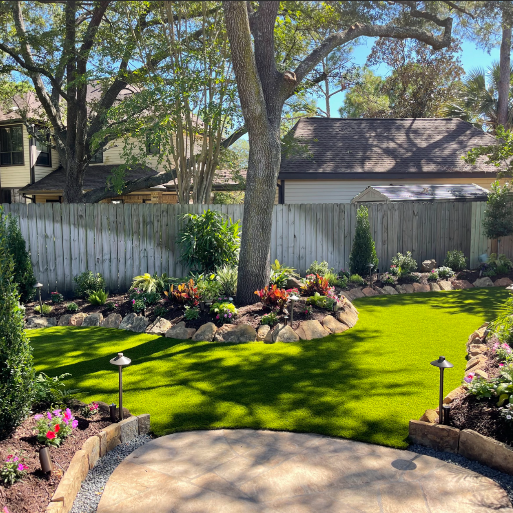 Backyard with artificial turf, detailed landscaping, and shaded area under large trees near Friendswood, TX.
