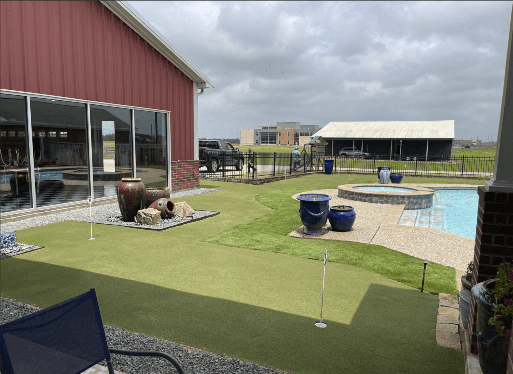 Custom backyard putting green installation surrounding a swimming pool, providing a varied and challenging putting experience, with decorative urns and gravel accents, installed by Sermons Turf Installs.
