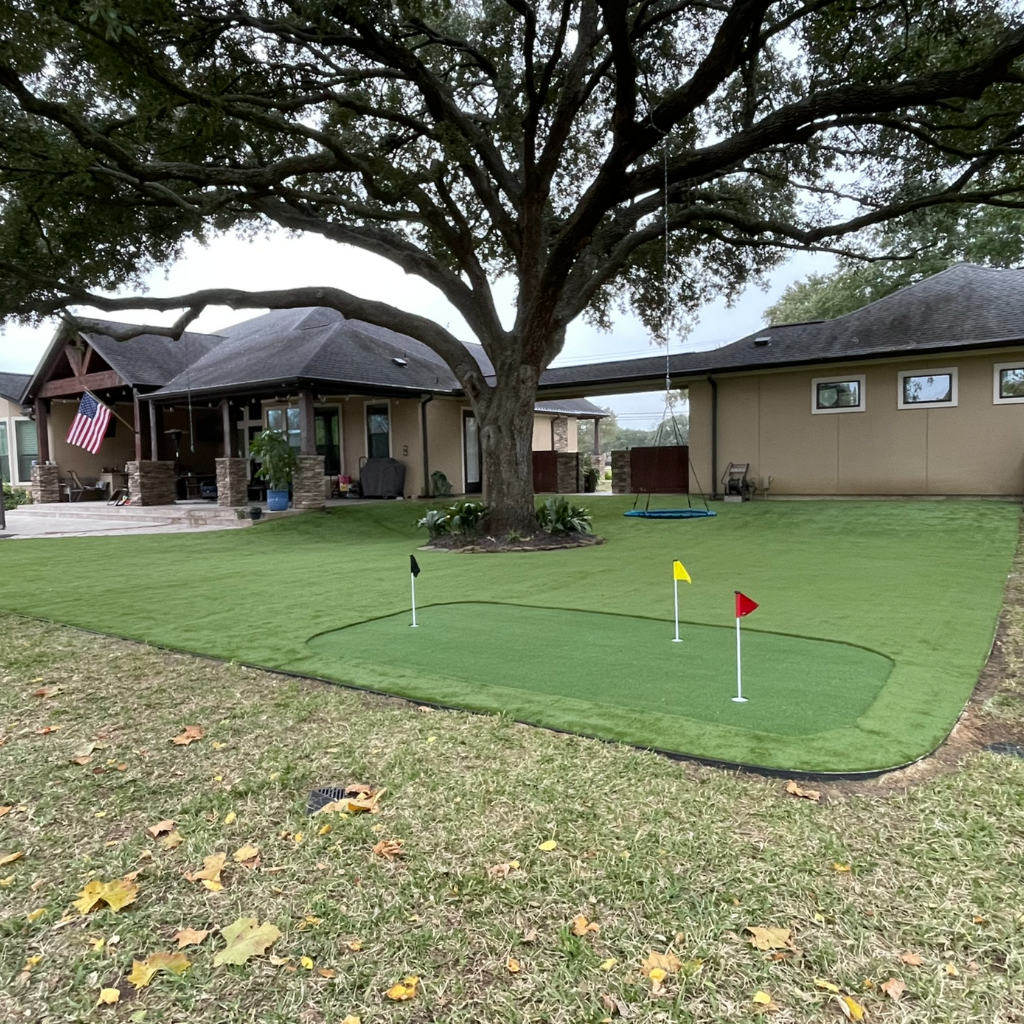 Custom backyard putting green installation with an adjacent patio, swing set, and shaded play area under a large oak tree, designed by Sermons Turf Installs.