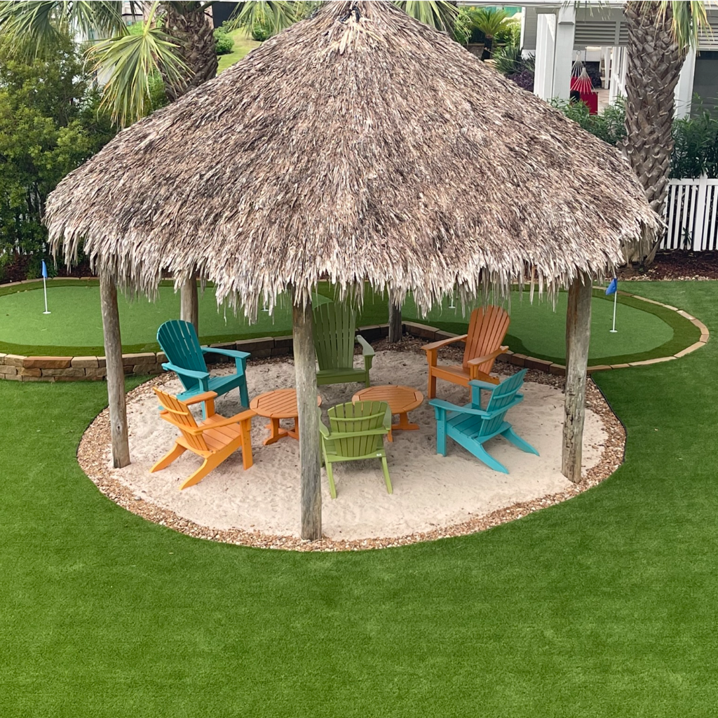 Aerial view of a waterfront putting green by Sermons Turf Installs near Galveston Bay, featuring a tiki hut with colorful Adirondack chairs.