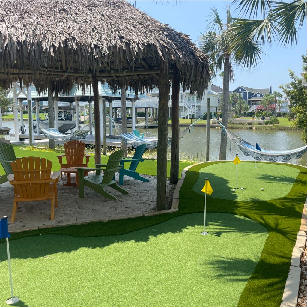 Waterfront putting green installation by Sermons Turf Installs near Galveston Bay, with a tiki hut, Adirondack chairs, and hammocks, surrounded by lush landscaping.