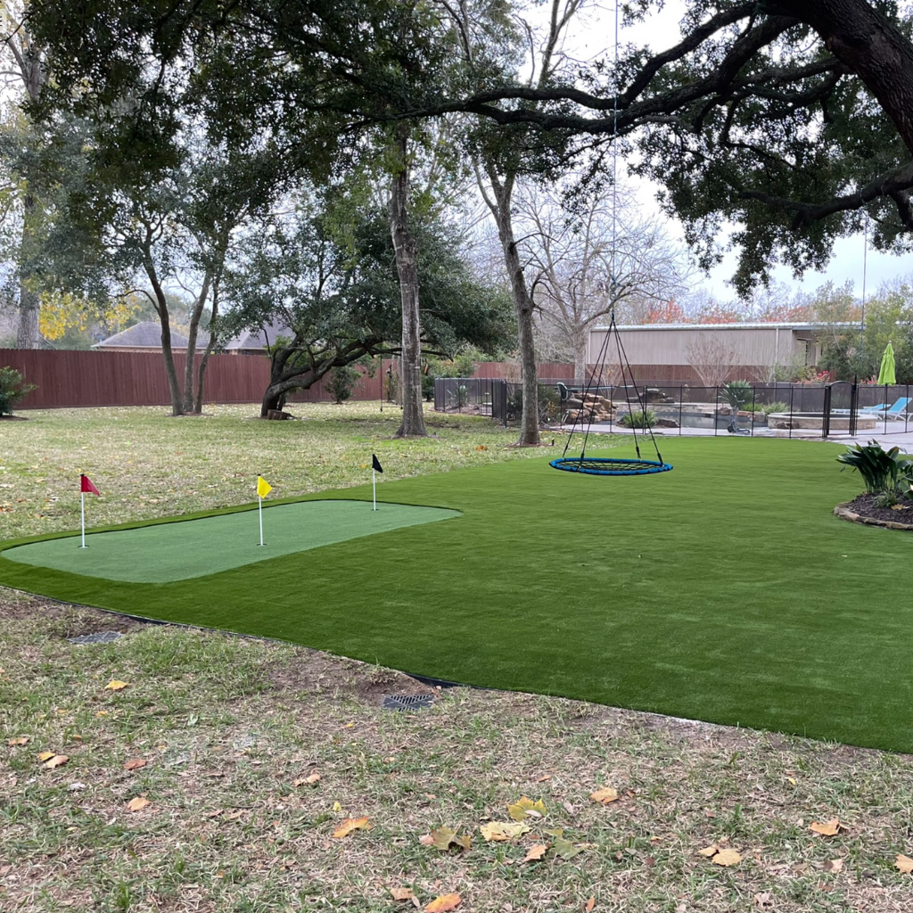 A backyard putting green installation in suburban South Houston, shaded by oak trees, with a nearby swing and swimming pool, by Sermons Turf Installs.